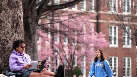 Student working on his laptop as another student walks by on Polk Place on the campus of UNC-Chapel Hill.